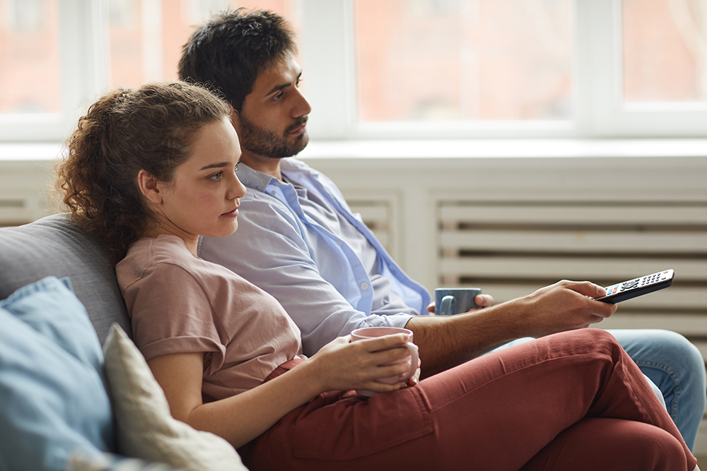 a side view portrait modern couple watching tv holding mugs while sitting sofa home cozy apartment