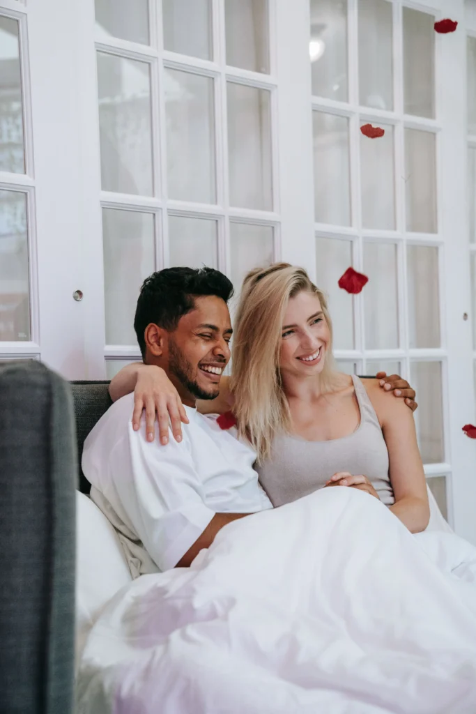 A smiling young man and blonde woman sit up in a white bed, laughing together as red rose petals fall around them in front of a glass paneled door.