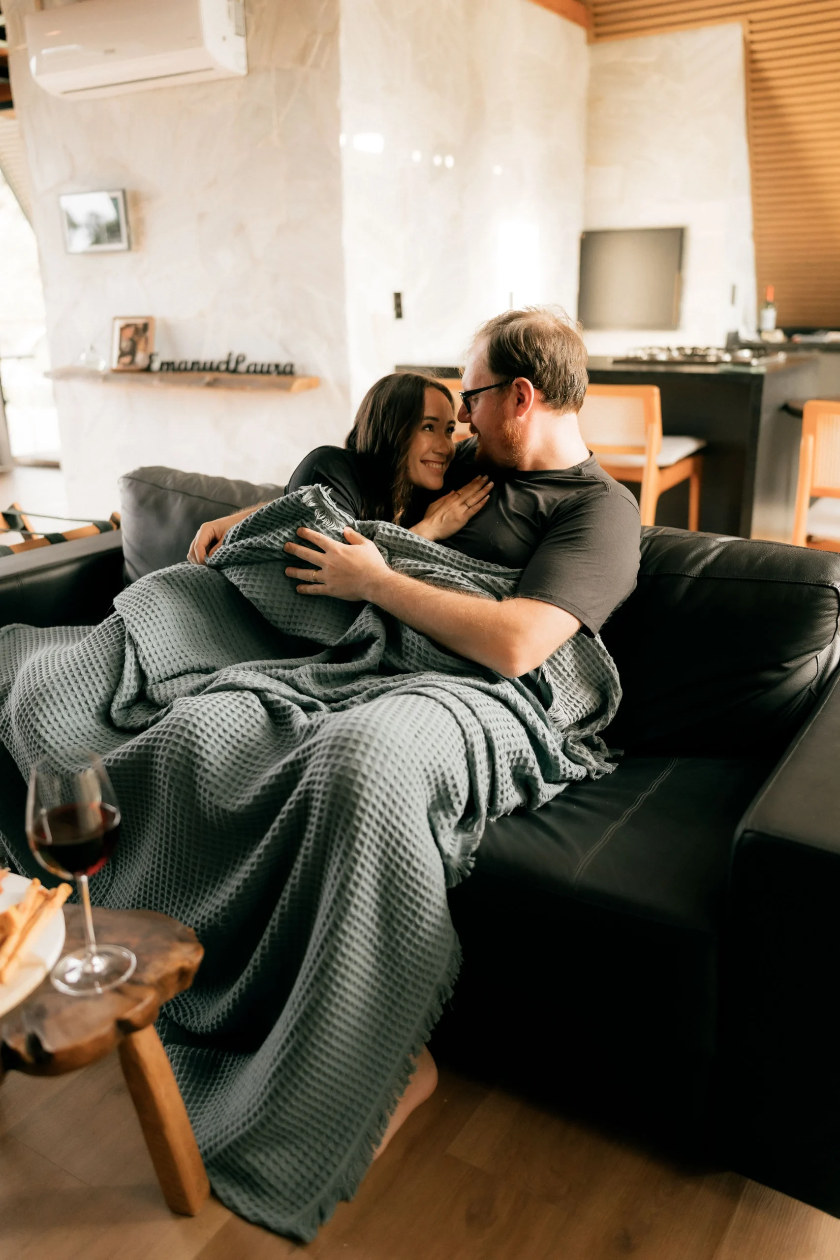 A happy couple cuddling on a black leather sofa under a grey waffle-weave blanket, sharing a warm romantic moment.