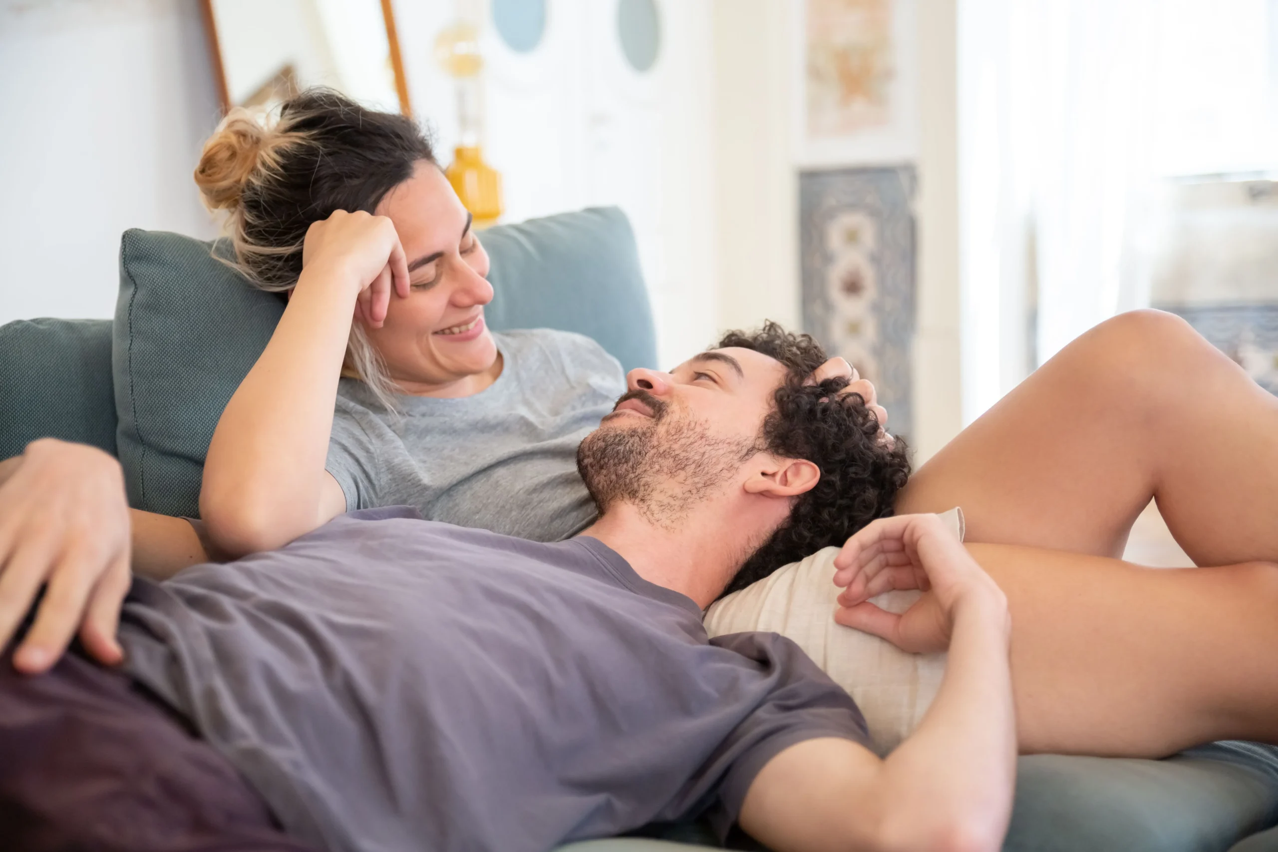 A smiling woman with her hair in a bun rests on a couch while gently running her fingers through her partner's curly hair as he lies in her lap.