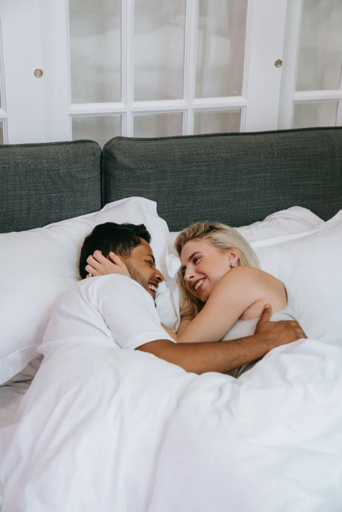 A happy, laughing young couple cuddling under warm white sheets in bed, looking lovingly at each other during a cozy morning.