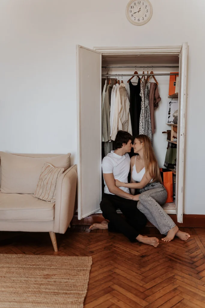A young romantic couple sitting barefoot on a wooden floor inside an open closet, leaning in to kiss amidst hanging clothes.