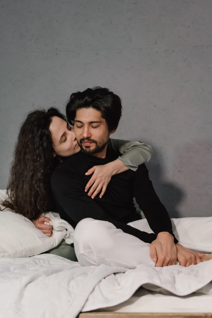 A woman with dark curly hair hugs a man in a black long-sleeved shirt from behind as they sit together closely on a bed with a neutral grey background