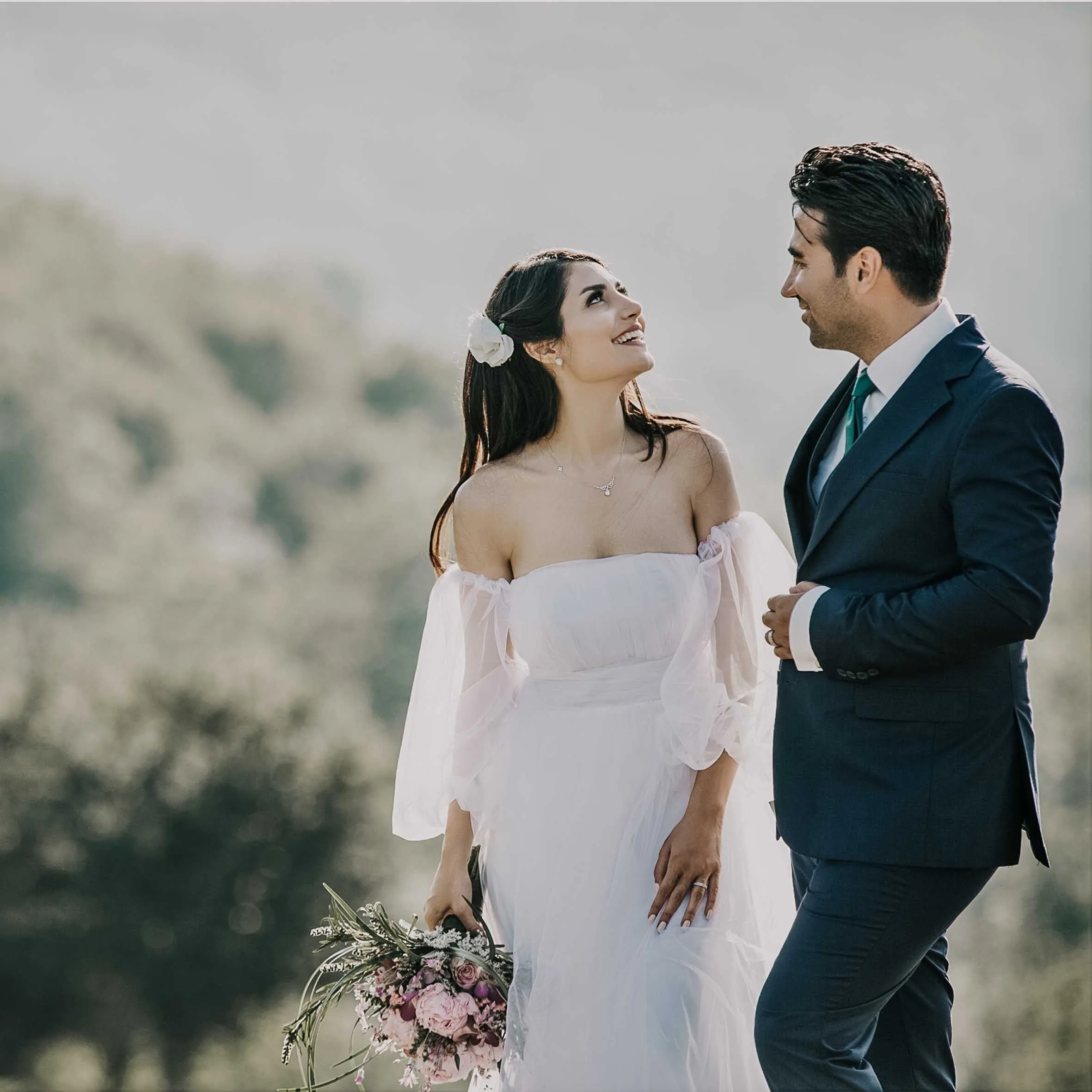 A bride holding a bouquet looking at her groom outdoors.