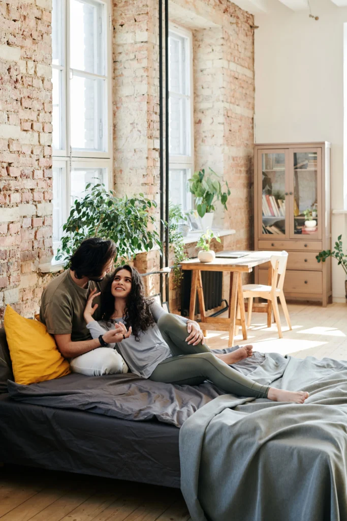 A young couple sitting together on a bed in a loft apartment with exposed brick walls.