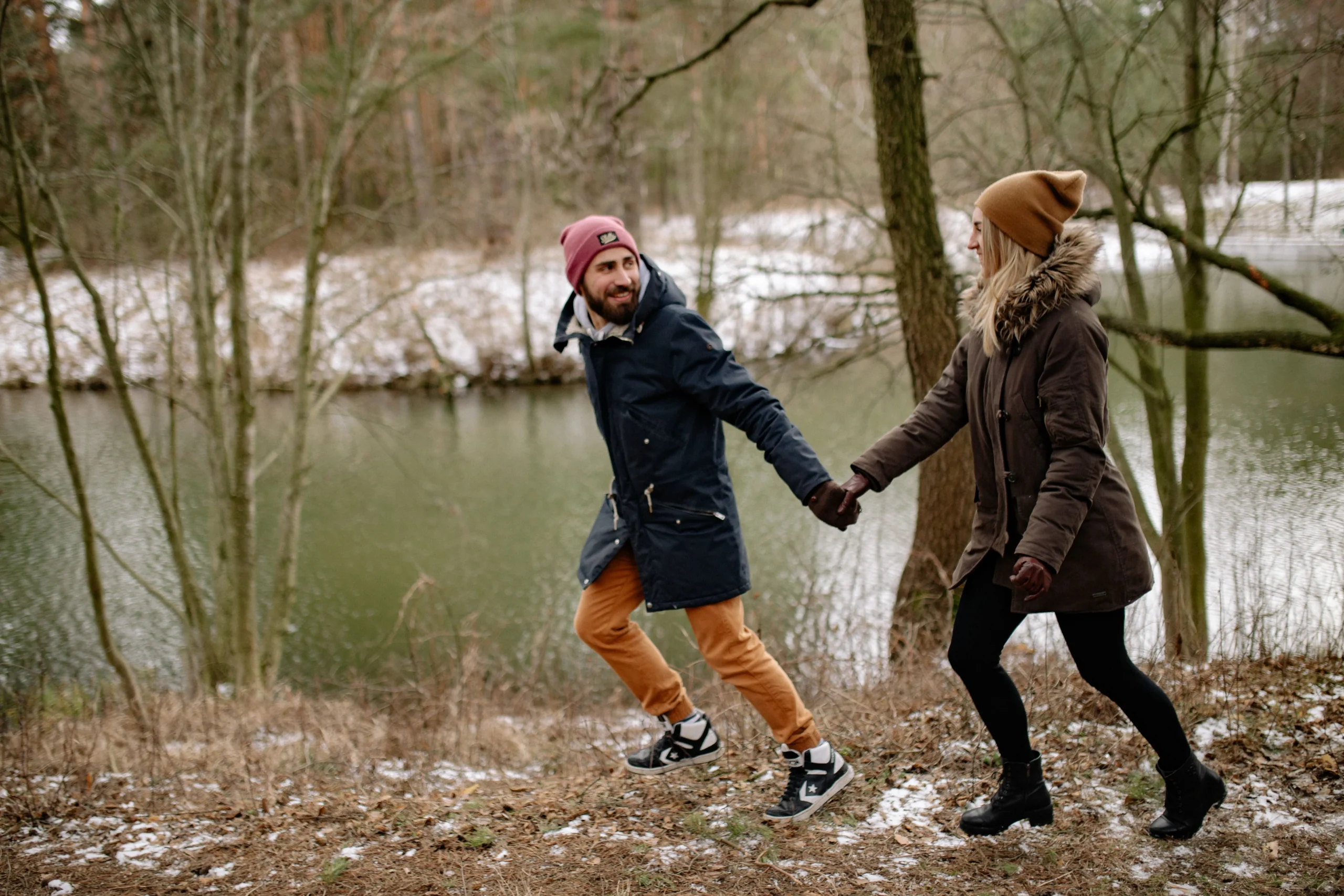 A man and woman holding hands while walking along a snowy path by a lake.