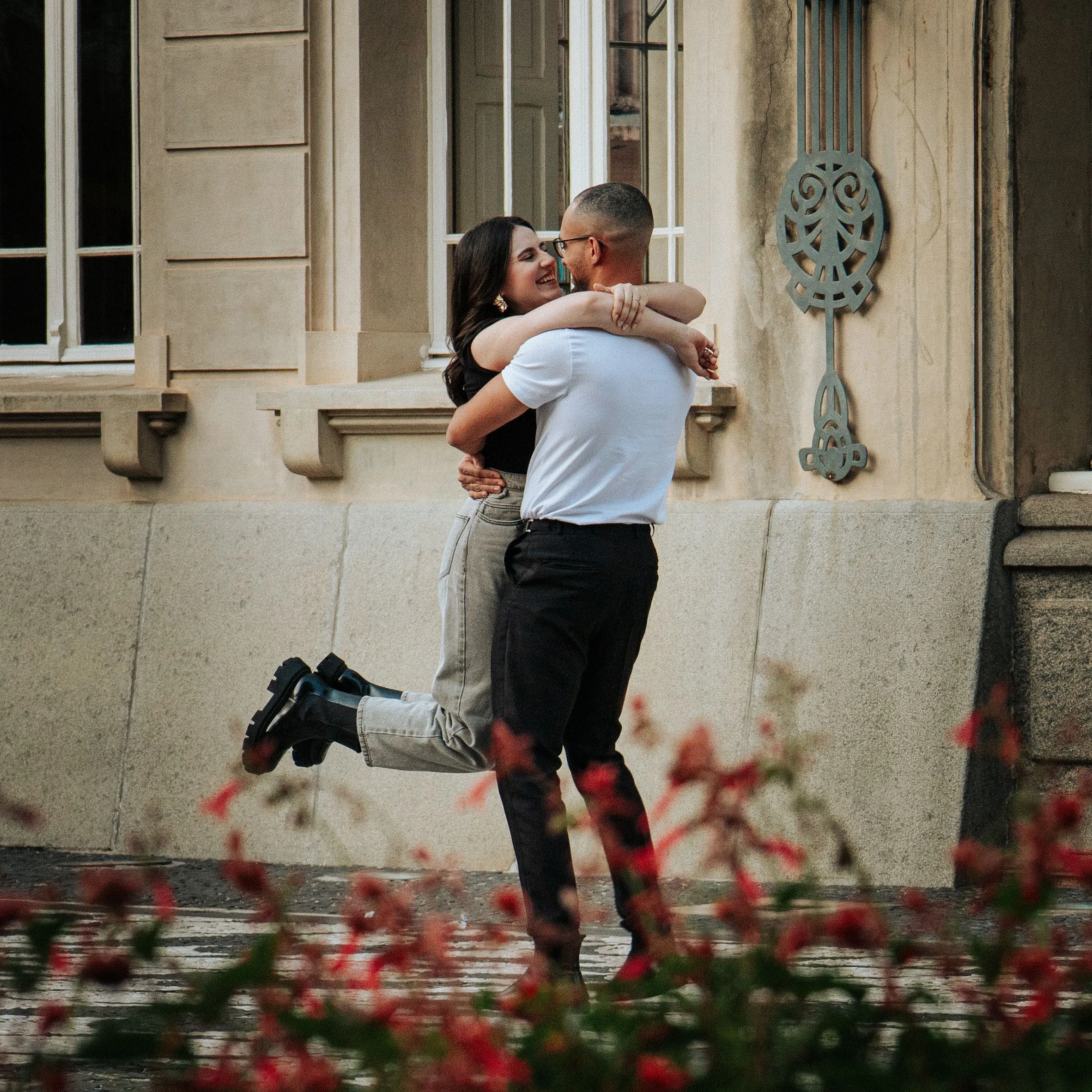 A man lifting and hugging a woman outside an old building with red flowers in the foreground.