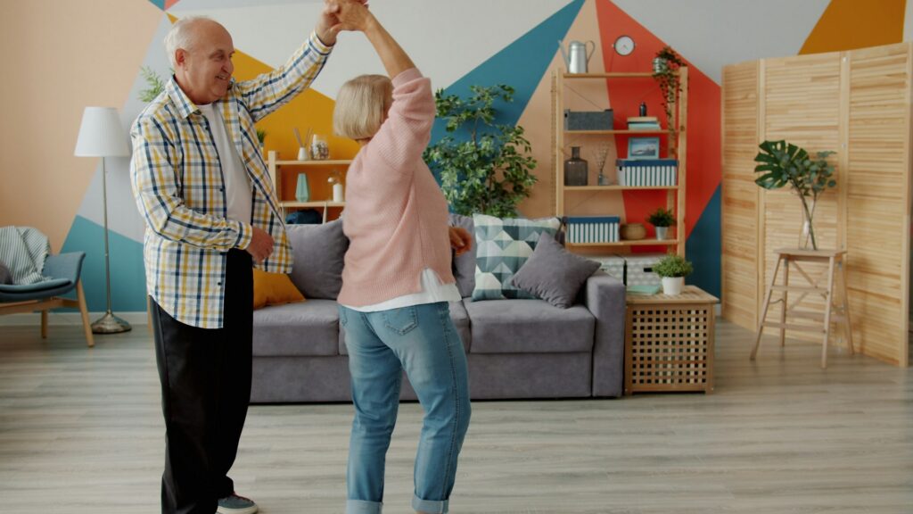 A cheerful older couple holding hands and dancing together in their living room at home